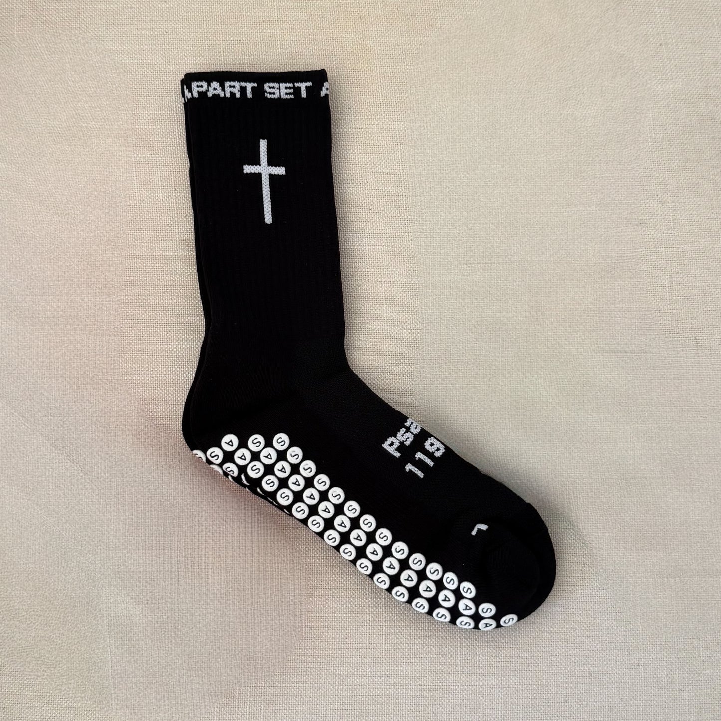 A single black crew sock with white printed graphics, including a cross on the calf, white text near the top, and a row of white smiley faces across the sole, laid flat on a beige textured fabric background.